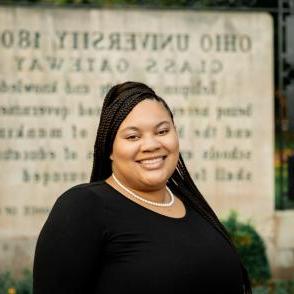 Darian Andrews smiles in front of the Ohio University Class Gateway, she wears a black shirt and a pearl necklace.